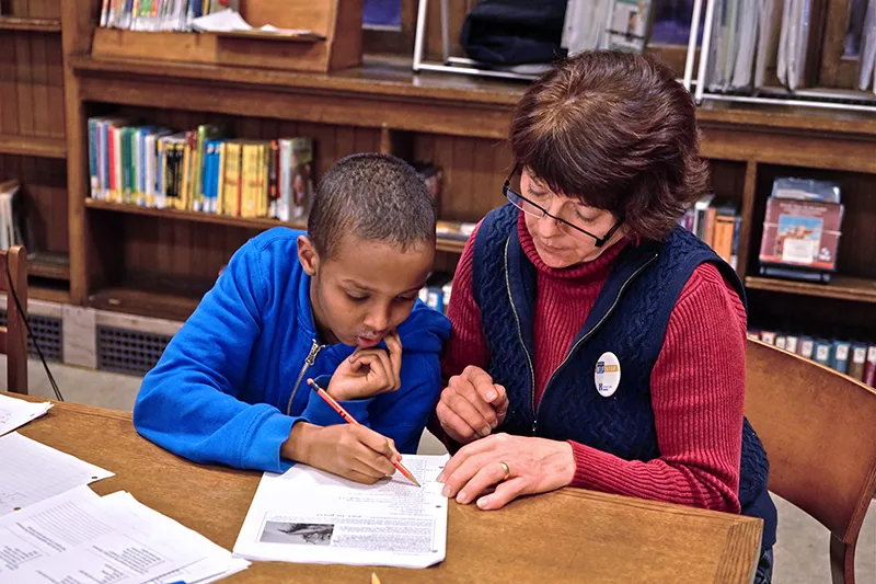 An adult helping a child with homework by the table surrounded by books