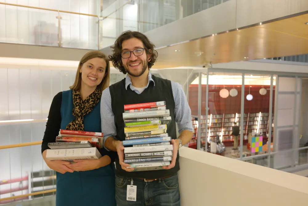 Two librarians stand at the stairwell of Minneapolis Central Library holding stacks of books.