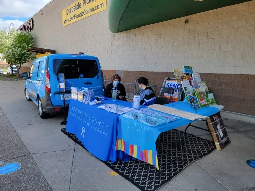 The library van is parked outside of a grocery story and staffed by two librarians