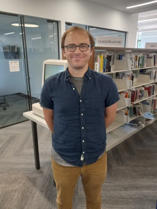 A person wearing glasses and smiling in front of a library book shelf