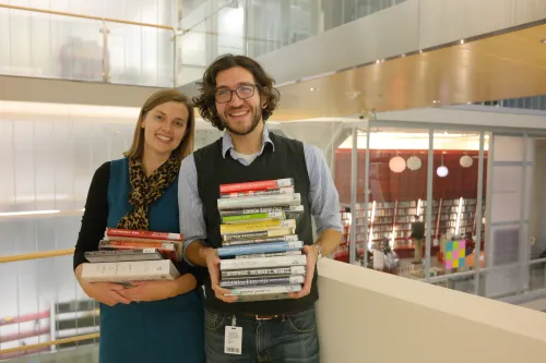 Two librarians stand at the stairwell of Minneapolis Central Library holding stacks of books.