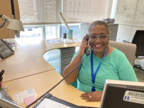 Gale Cannon, Friends development associate, sits at a desk smiling with her ear to a telephone. She wears polka dot glasses and a bright, cheerful shirt,