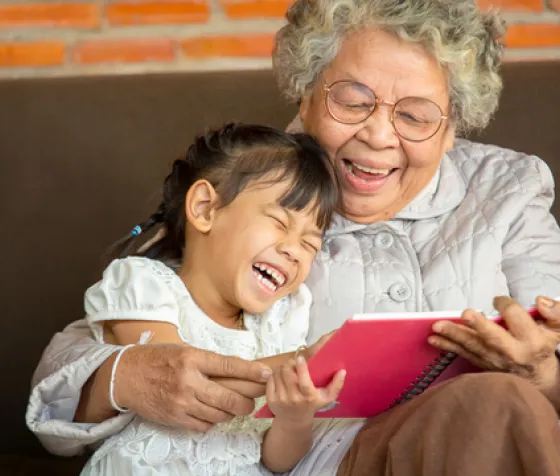 Grandma and granddaughter laughing while sitting and reading a book together 