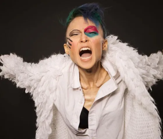 Mayda wears dramatic, bright makeup and sings passionately on a dark stage.