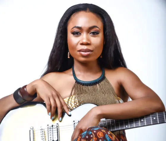 Carolyne sits in front of a white wall, holding her silver guitar and wearing a large beaded necklace.