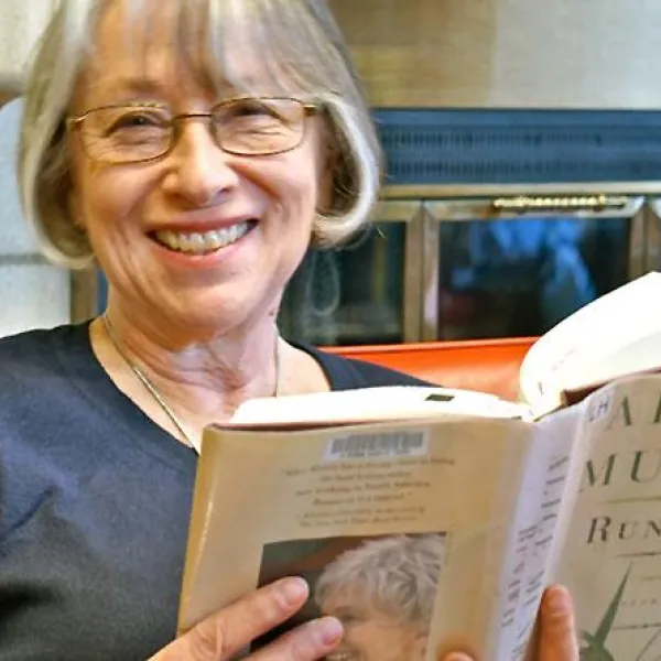 Older woman wearing glasses is smiling while reading book by the fireplace